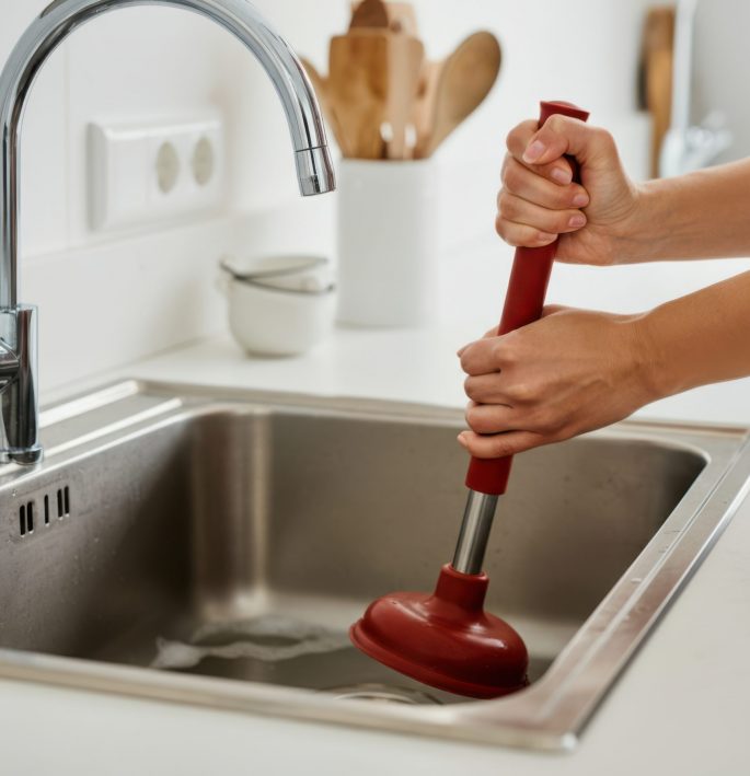 Person unclogs a kitchen sink using a plunger in a modern kitchen with minimalist decor during daylight